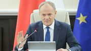 Mężczyzna w garniturze siedzi przy stole, gestykulując, z flagami w tle. Donald Tusk.