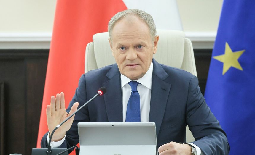 Mężczyzna w garniturze siedzi przy stole, gestykulując, z flagami w tle. Donald Tusk.