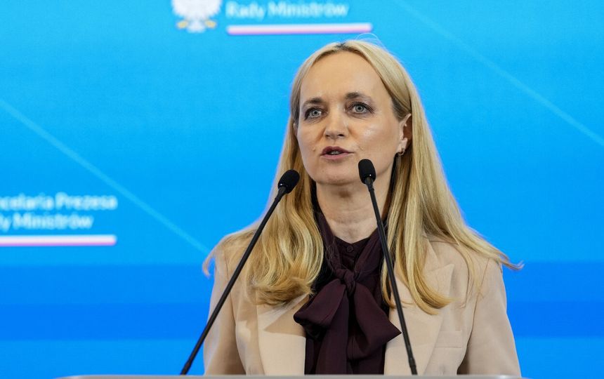 Kobieta z długimi blond włosami przemawia przy mównicy, na tle niebieskiego tła z logo.