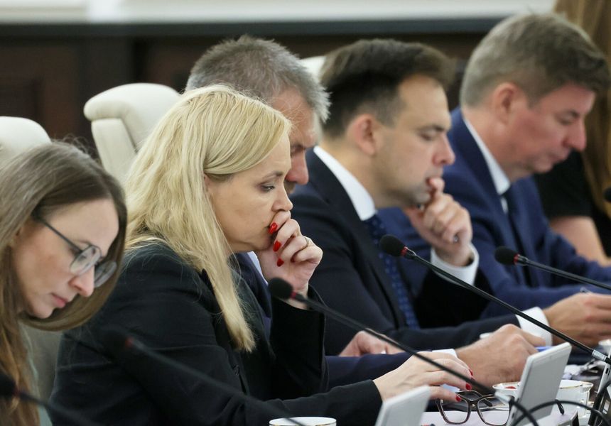 Izba dyskusyjna, sześć osób w garniturach, skupionych nad dokumentami. Kobieta z blond włosami na pierwszym planie.