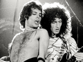 Freddie Mercury i Brian May. Na zdjęciu dwóch mężczyzn gra na gitarach podczas koncertu, w tle ciemne światła.