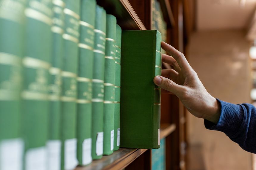 Osoba sięga po zieloną książkę na półce w bibliotece. W tle widać inne podobne tomy.