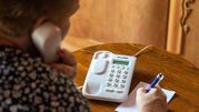 Kobieta rozmawia przez telefon stacjonarny, pisząc coś na kartce. Drewniany stół w tle.