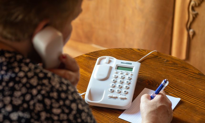Kobieta rozmawia przez telefon stacjonarny, pisząc coś na kartce. Drewniany stół w tle.