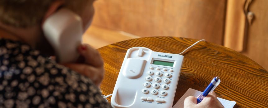 Kobieta rozmawia przez telefon stacjonarny, pisząc coś na kartce. Drewniany stół w tle.