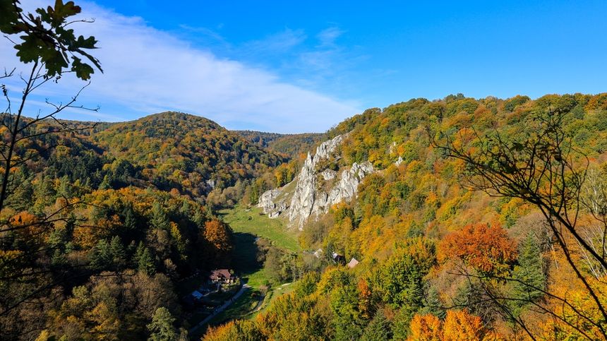 Górski krajobraz w jesiennych barwach, z zielonymi i pomarańczowymi drzewami oraz błękitnym niebem.