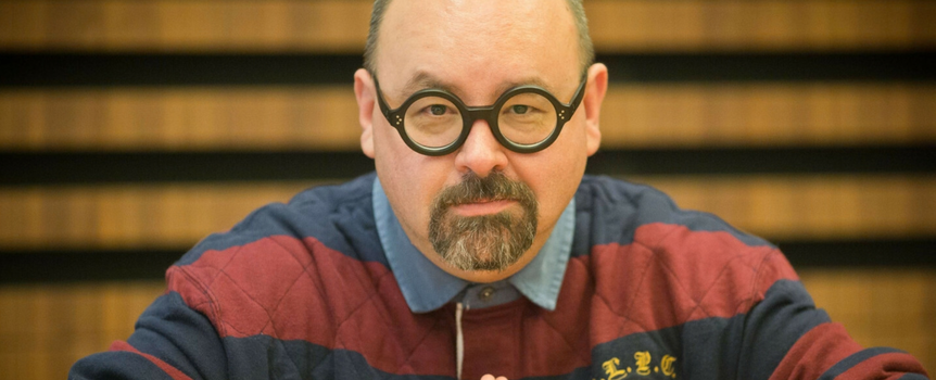 Mężczyzna w okrągłych okularach, w koszulce w paski, przy stole z poważnym wyrazem twarzy.