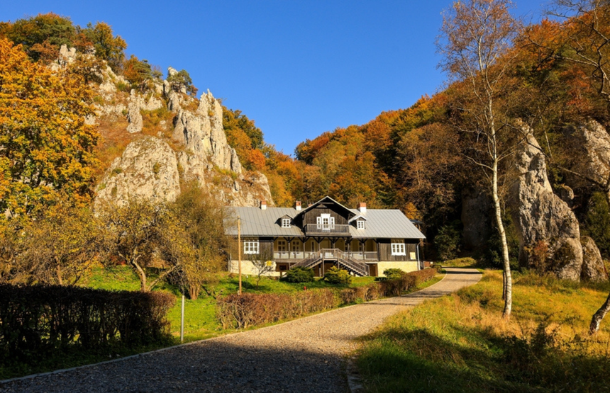 Widok na drewniany dom wśród kolorowych drzew i skalnych formacji pod niebieskim niebem.