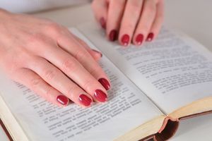 Na zdjęciu widoczne są ręce z czerwonymi paznokciami spoczywające na otwartej książce.