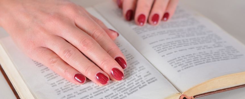 Na zdjęciu widoczne są ręce z czerwonymi paznokciami spoczywające na otwartej książce.