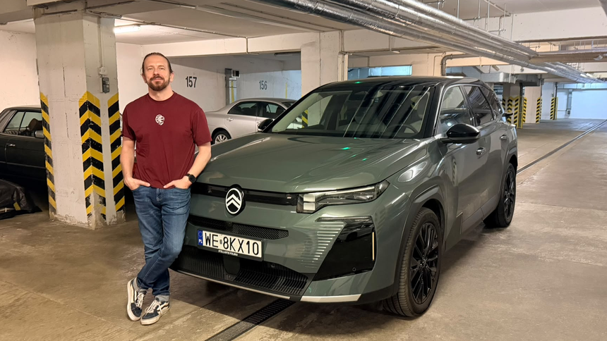 Mężczyzna w t-shircie stoi obok zielonego SUV-a w garażu. Tło z innymi samochodami.