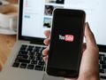 Smartfon trzymany w ręce, na wyświetlaczu logo YouTube. W tle laptop i filiżanka kawy.