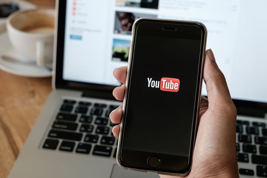 Smartfon trzymany w ręce, na wyświetlaczu logo YouTube. W tle laptop i filiżanka kawy.