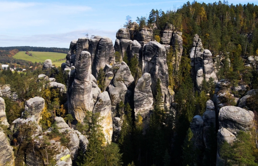 Widok na skalne formacje otoczone lasem, z zielonymi łąkami w tle. Słoneczny dzień.