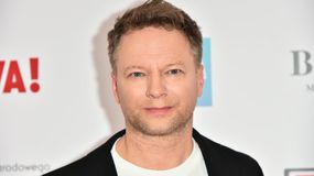 Maciej Stuhr z krótkimi, jasnymi włosami, uśmiecha się w czarnym żakiecie na jasnym tle.