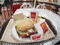 Na tacy znajduje się hamburger z sezamową bułką, frytki i napój, w tle stół w McDonald's.