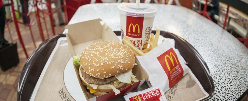 Na tacy znajduje się hamburger z sezamową bułką, frytki i napój, w tle stół w McDonald's.