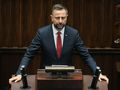 Na zdjęciu mężczyzna w ciemnym garniturze z czerwonym krawatem w parlamencie.