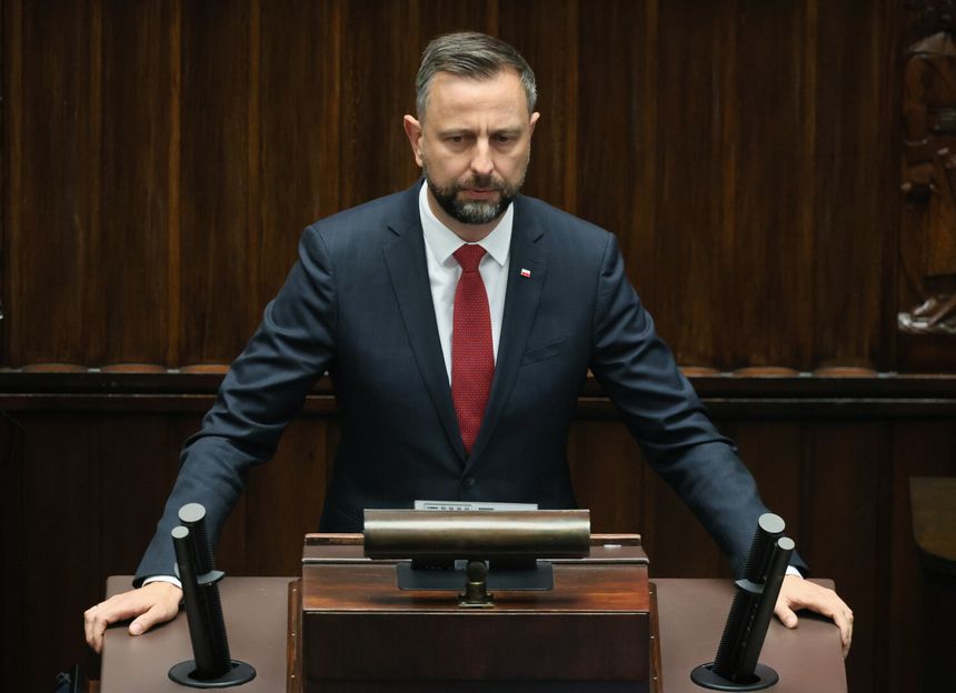 Na zdjęciu mężczyzna w ciemnym garniturze z czerwonym krawatem w parlamencie.