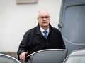 Mężczyzna w ciemnym płaszczu stoi obok samochodu, z poważnym wyrazem twarzy.
