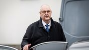 Mężczyzna w ciemnym płaszczu stoi obok samochodu, z poważnym wyrazem twarzy.