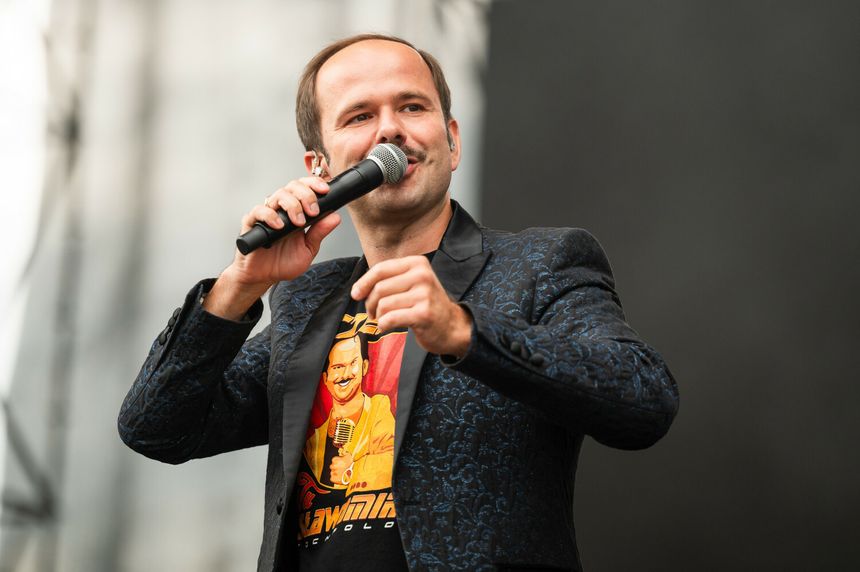 Mężczyzna w eleganckiej marynarce i t-shircie, trzyma mikrofon i uśmiecha się do tłumu.