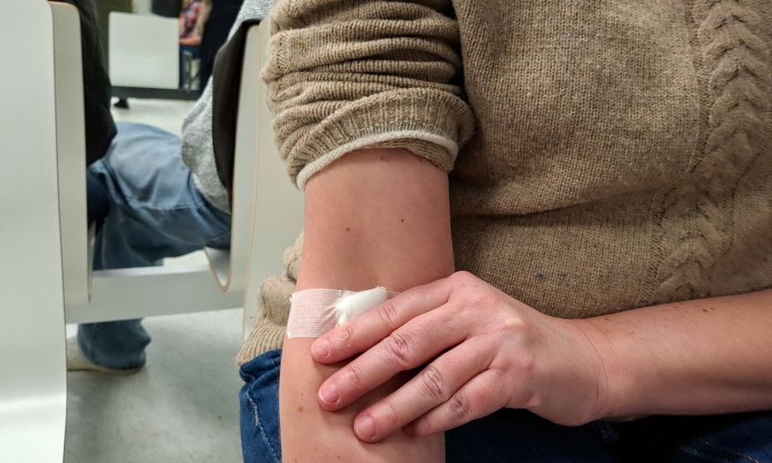 Na zdjęciu widoczna jest ręka z bandażem, osoba dotyka bandażu, w tle inni ludzie.