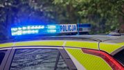 Migająca niebieska sygnalizacja na dachu radiowozu policyjnego, w tle zieleń drzew.