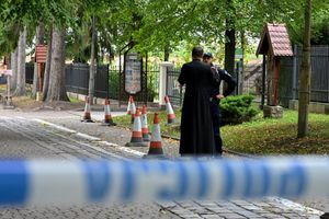 Dwóch mężczyzn rozmawia przy policyjnych pachołkach na ulicy, w tle drzewa i ogrodzenie.