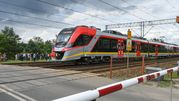 Pociąg przejeżdża przez przejazd kolejowy, a w tle grupka ludzi i znaki ostrzegawcze.