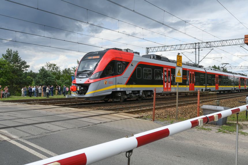 Pociąg przejeżdża przez przejazd kolejowy, a w tle grupka ludzi i znaki ostrzegawcze.