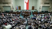 Posiedzenie Sejmu, liczni posłowie w sali, na ścianie biało-czerwona flaga i godło Polski.