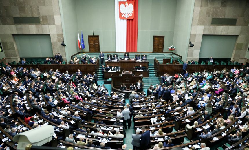Posiedzenie Sejmu, liczni posłowie w sali, na ścianie biało-czerwona flaga i godło Polski.