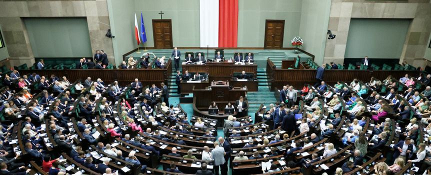 Posiedzenie Sejmu, liczni posłowie w sali, na ścianie biało-czerwona flaga i godło Polski.