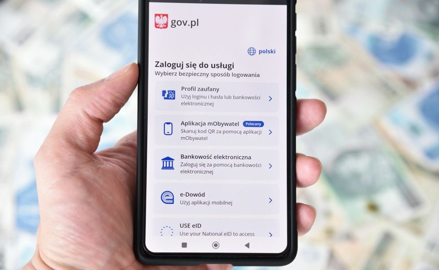 Smartfon w dłoni z ekranem logowania do usług e-administracji w Polsce. Tło to pieniądze.