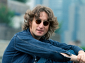 John Lennon w 1974 roku. Mężczyzna w okularach przeciwsłonecznych, ubrany w niebieską kurtkę, siedzi na tle miejskiego krajobrazu.