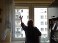 Kobieta w kratkowanej koszuli myje okno w jasnym pomieszczeniu z widokiem na bloki.