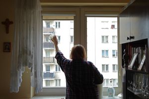 Kobieta w kratkowanej koszuli myje okno w jasnym pomieszczeniu z widokiem na bloki.