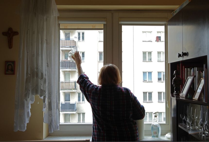 Kobieta w kratkowanej koszuli myje okno w jasnym pomieszczeniu z widokiem na bloki.