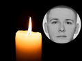 Świeca zapalona na czarnym tle, obok okrągłe zdjęcie mężczyzny z poważnym wyrazem twarzy.