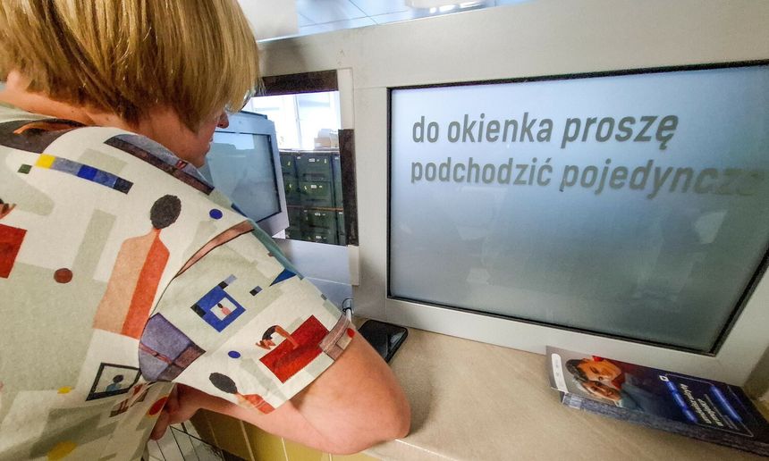 Osoba o krótkich włosach podchodzi do okienka z wyświetlaczem z napisem.