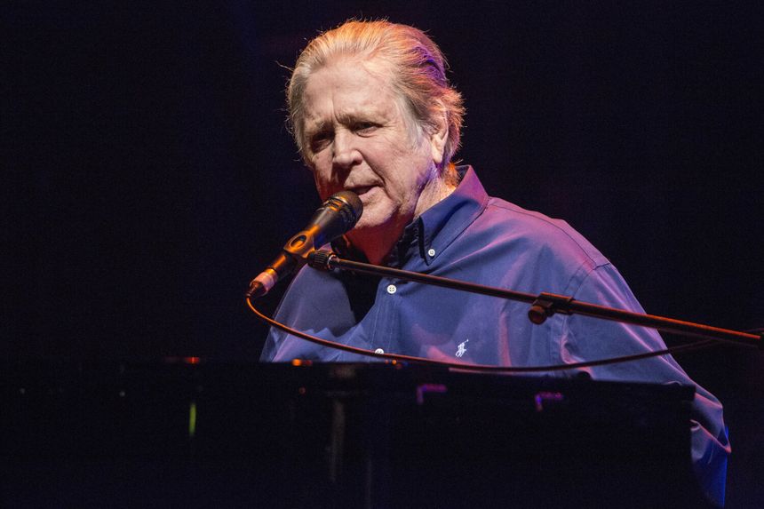 Brian Wilson w 2016 roku. Mężczyzna w niebieskiej koszuli śpiewa do mikrofonu, stoi przy fortepianie na scenie.
