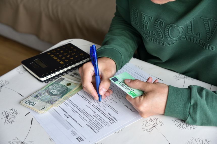 Osoba w zielonej bluzie wypełnia formularz, obok leżą banknoty i kalkulator.