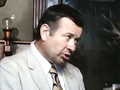 Mężczyzna w jasnym garniturze, z krótkimi włosami, mówi w pomieszczeniu z drewnianymi akcentami.
