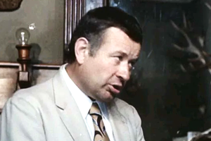 Mężczyzna w jasnym garniturze, z krótkimi włosami, mówi w pomieszczeniu z drewnianymi akcentami.