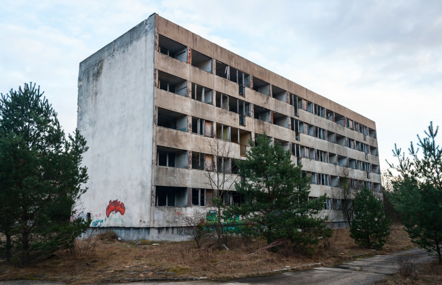 Opuszczony budynek wielorodzinny z widocznymi zniszczeniami i rosnącą roślinnością wokół.