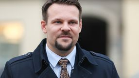 Mężczyzna w ciemnym płaszczu z krawatem, uśmiechający się w miejskim otoczeniu.