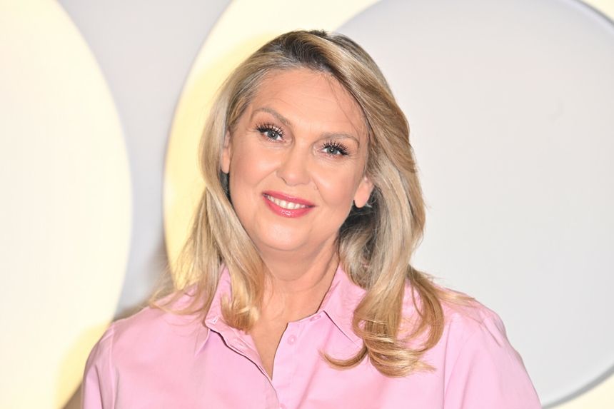 Kobieta z długimi, blond włosami w różowej koszuli, uśmiecha się do kamery.