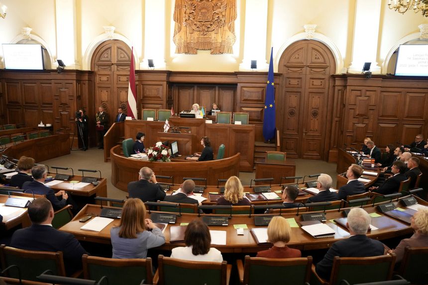 Zgromadzenie w parlamencie, mężczyzna przy mównicy, kobiety siedzące, flagi w tle.