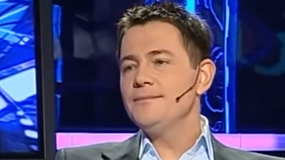 Krzysztof Ibisz z mikrofonem przedstawiony na tle ekranów, uśmiechający się podczas rozmowy.
